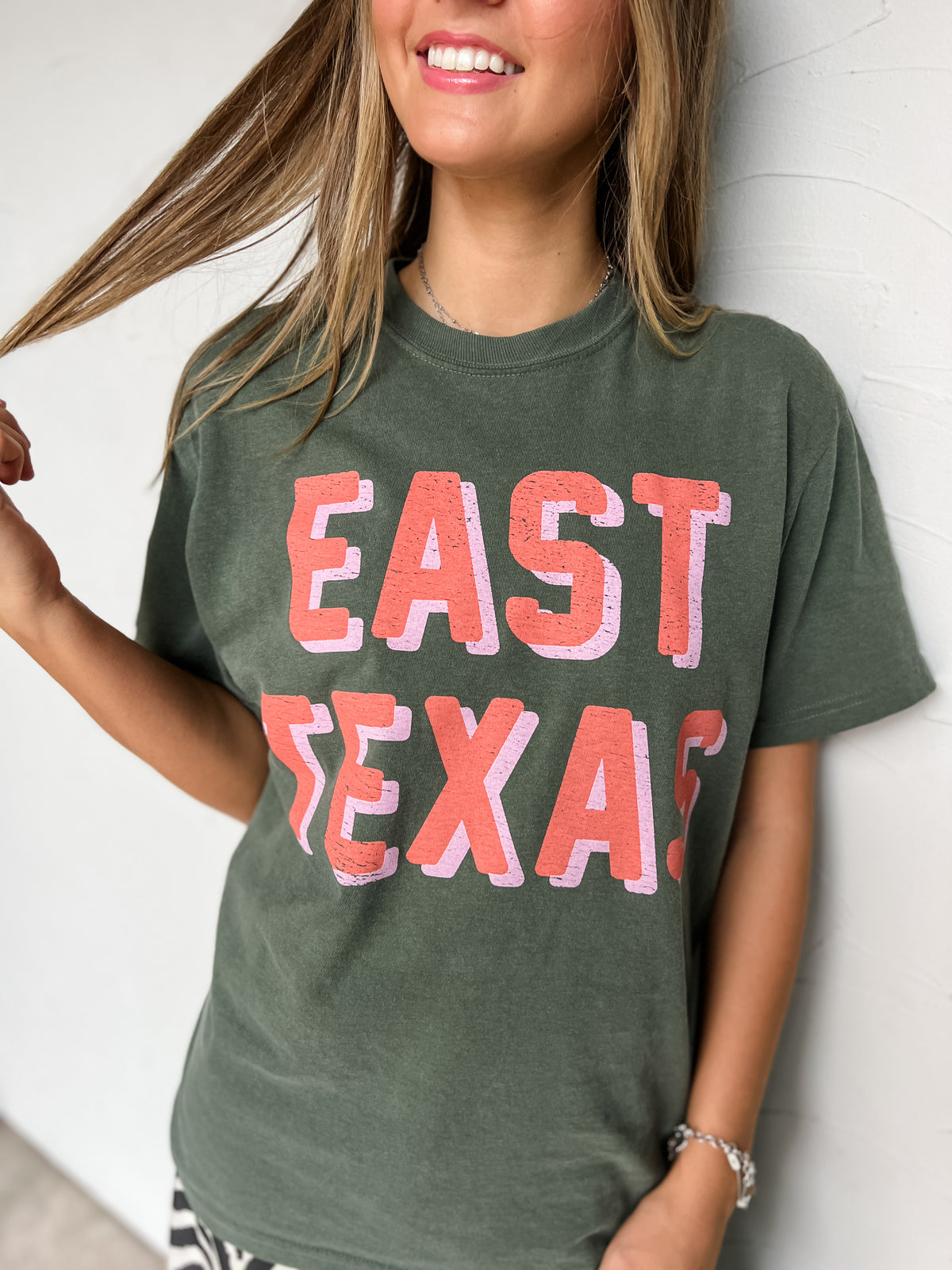 A woman wearing a faded olive green graphic t-shirt with 'EAST TEXAS' printed in distressed coral pink letters with a light pink drop shadow, leaning against a white wall.