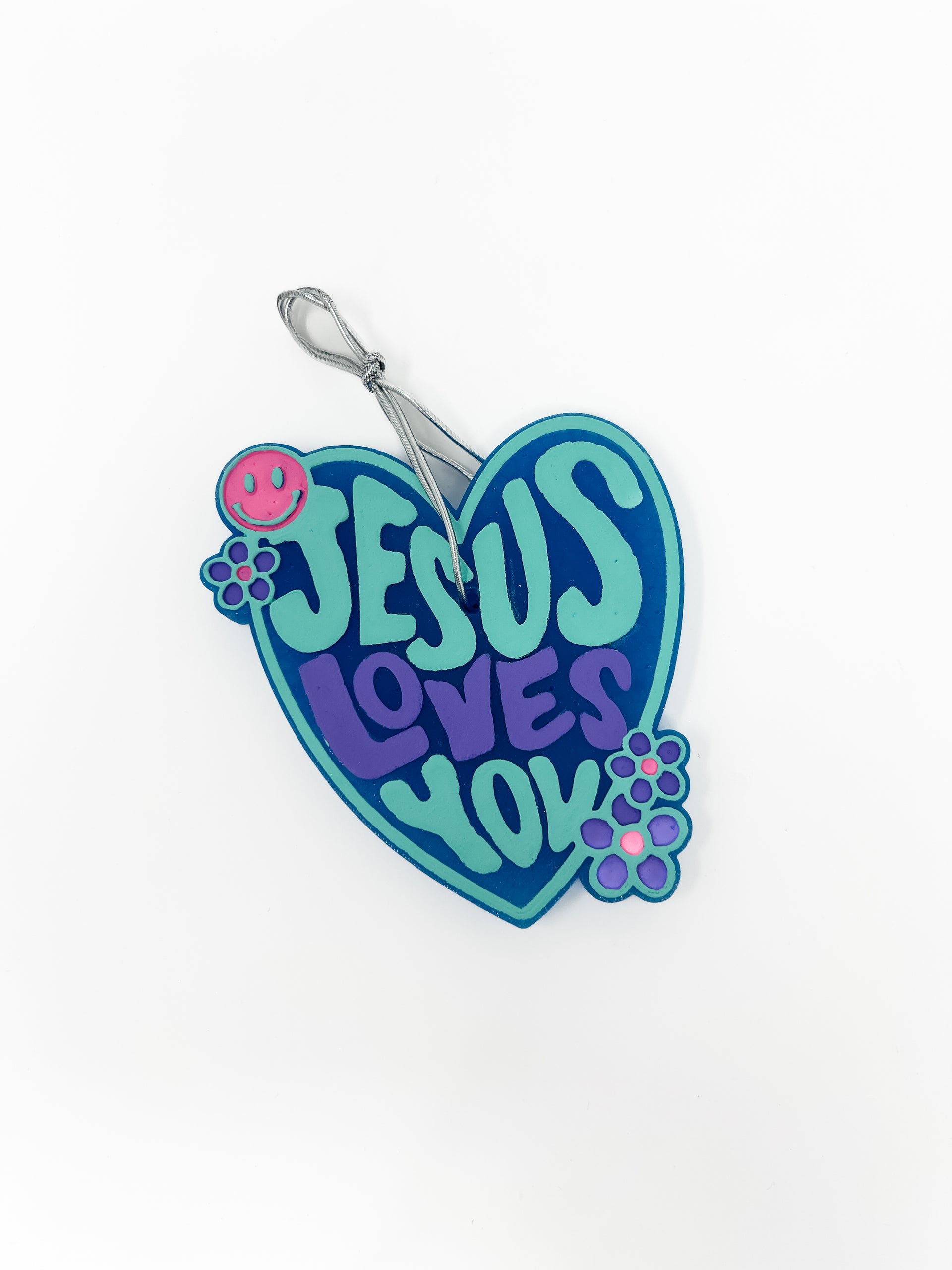 This "Jesus Loves You" car freshie features a groovy, heart-shaped design with retro lettering, accented by a tiny smiley face and small flower details along the border. It comes ready to hang from your rearview mirror with an attached hanging hook. Colors and scents vary.