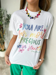 A person wearing a white graphic T-shirt with colorful hand-lettered text reading 'YOU ARE FOR MORE PRECIOUS THAN JEWELS' and small floral/butterfly/gem accents, paired with bright green pants and a multicolor beaded necklace.
