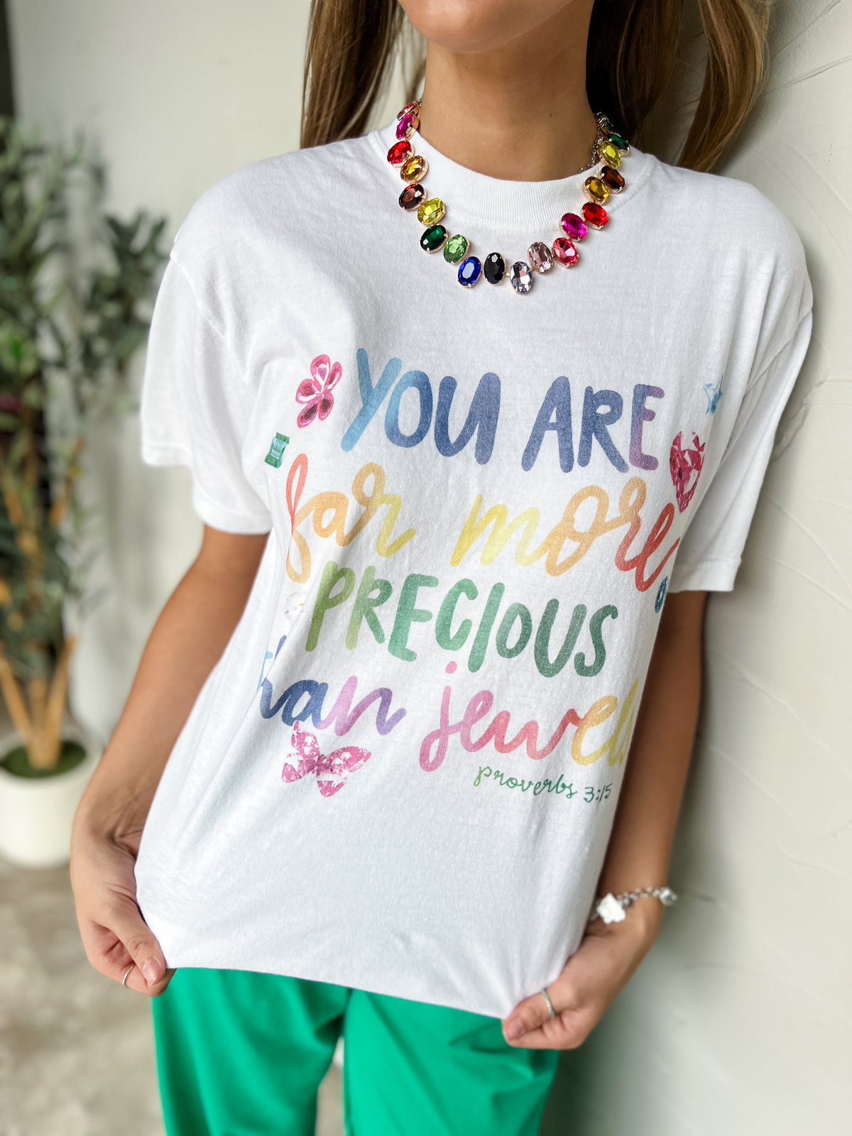 A person wearing a white graphic T-shirt with colorful hand-lettered text reading 'YOU ARE FOR MORE PRECIOUS THAN JEWELS' and small floral/butterfly/gem accents, paired with bright green pants and a multicolor beaded necklace.