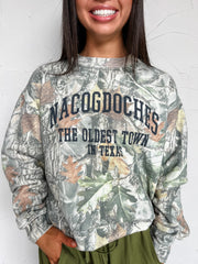 Woodland camo crewneck sweatshirt with “Nacogdoches, The Oldest Town in Texas” lettering, long sleeves, and banded cuffs and hem, made of 47% rayon, 46% polyester, and 7% spandex.
