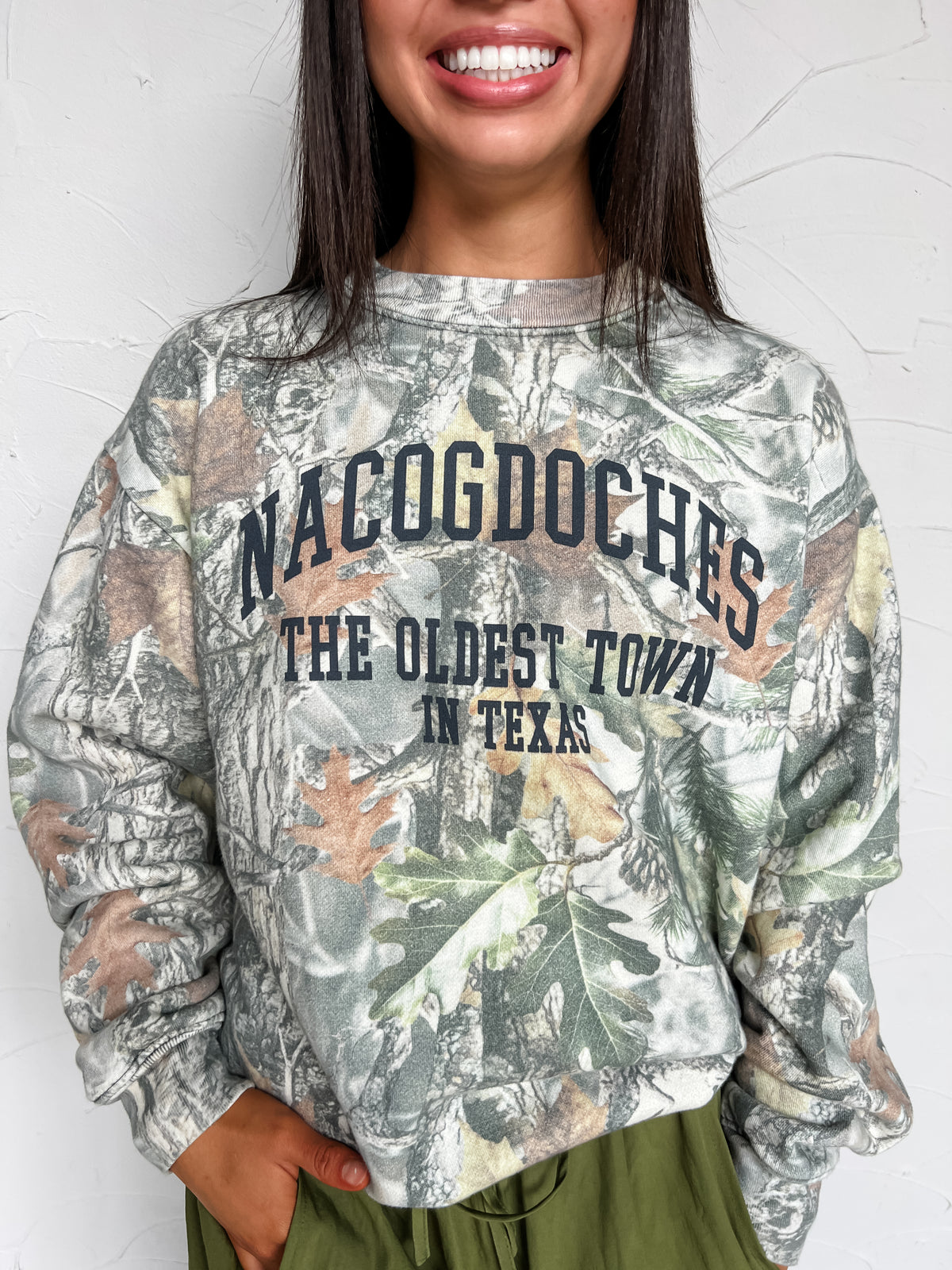 Woodland camo crewneck sweatshirt with “Nacogdoches, The Oldest Town in Texas” lettering, long sleeves, and banded cuffs and hem, made of 47% rayon, 46% polyester, and 7% spandex.
