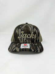 A Richardson 112PFP timber camo trucker hat embroidered with "Jacks" in raised 3D beige script thread, featuring tan mesh back panels, displayed against a solid white background.