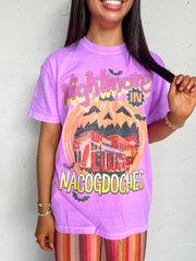 Purple Comfort Colors tee made of 100% garment-dyed cotton, featuring a “Nightmare In Nacogdoches” screenprint with a jack-o’-lantern, bats, and a downtown storefront scene.