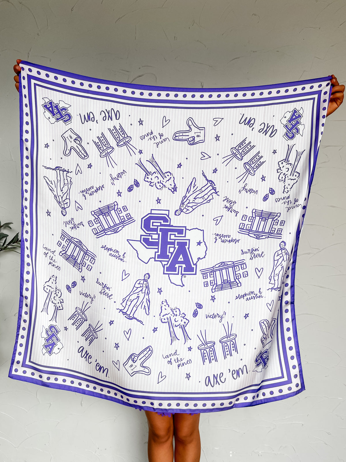 A white square scarf or bandana with a purple border and purple printed illustrations, held up by a person against a light gray wall. The design features the letters 'SFA' in a purple outline shape resembling Texas, surrounded by small drawings of hands, hearts, stars, and text reading 'One Team'.