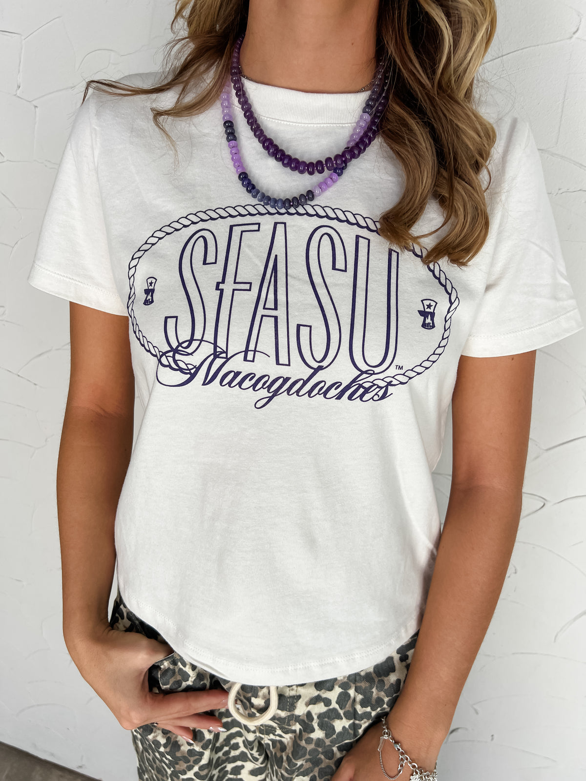 Model wearing the SFASU Vintage Rope white Bella Canvas 100% cotton unisex tee with purple screenprinted SFASU lettering, an oval rope border, and Nacogdoches script, styled with layered purple necklaces and leopard-print pants.