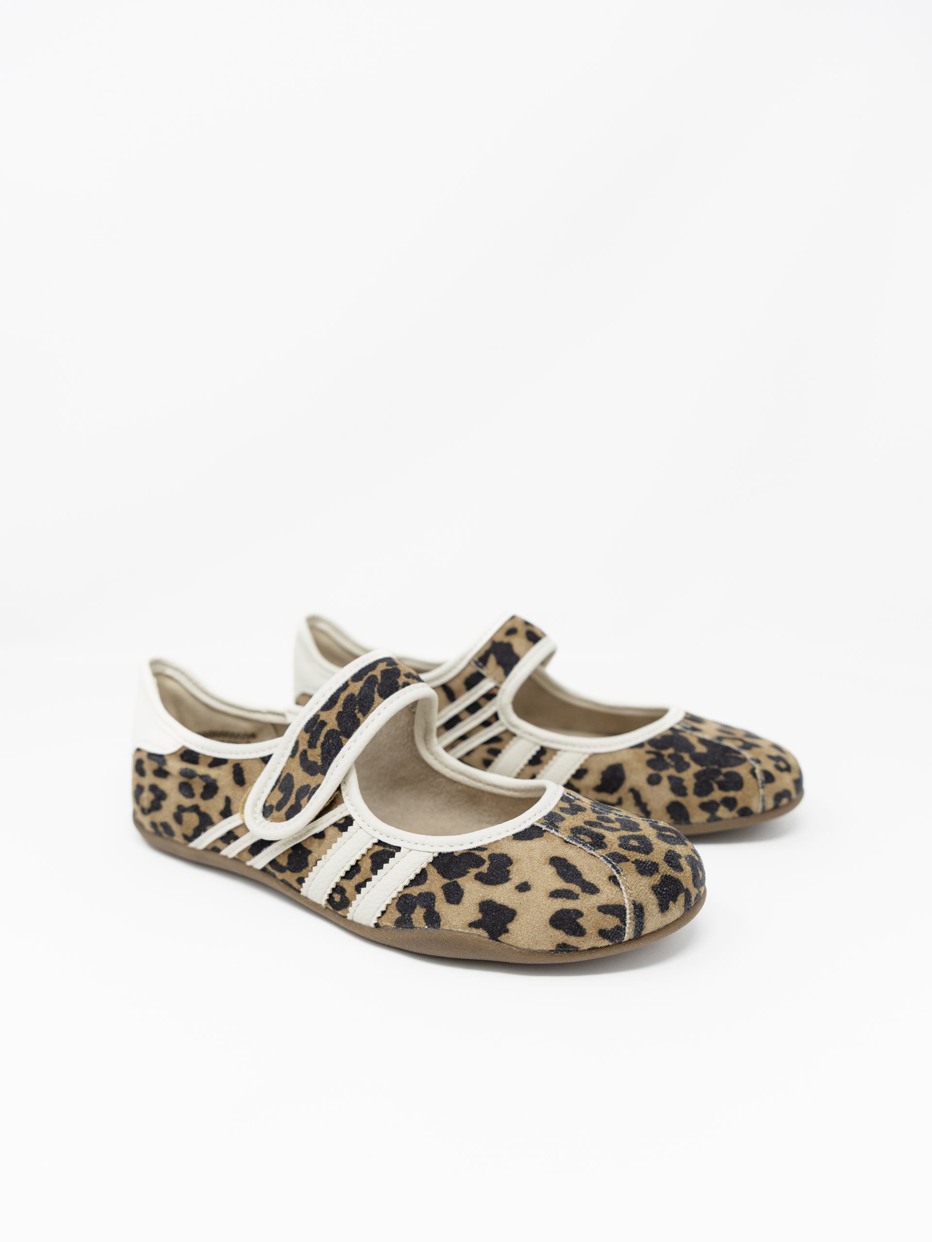 A pair of leopard print Mary Jane style flats with off-white trim and athletic stripes on the sides, set against a plain white background. The shoes feature an adjustable hook-and-loop strap across the top and a thin, flexible low-profile sole.