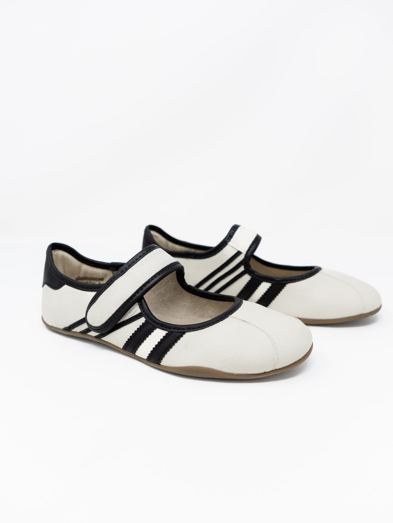 A pair of off-white pebbled faux leather Mary Jane style flats with black piping trim and athletic stripes on the sides, set against a solid white background. The shoes feature an adjustable hook-and-loop strap across the top and a thin, flexible low-profile sole.