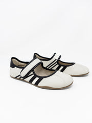 A pair of off-white pebbled faux leather Mary Jane style flats with black piping trim and athletic stripes on the sides, set against a solid white background. The shoes feature an adjustable hook-and-loop strap across the top and a thin, flexible low-profile sole.