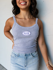 A mid-shot photograph focusing from the waist to the mouth of a smiling young woman with dark wavy hair posing with one hand on her hip against a textured white wall. She is wearing a fitted heather grey ribbed tank top made from 100% cotton fabric, featuring a scoop neckline and an oval purple embroidered patch centered at the chest reading "SFA", paired with a light wash denim skirt and stacked gold and pearl bracelets.