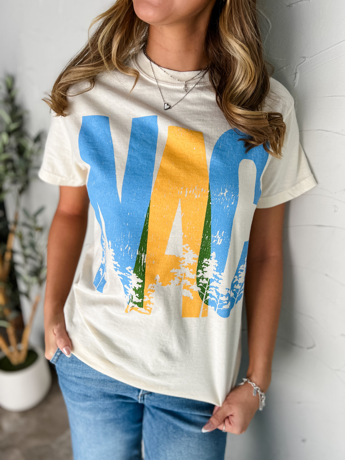 Catie wearing the NAC Overlap ivory Comfort Colors 100% cotton tee in size Small, featuring oversized overlapping blue and golden-yellow NAC lettering, green accents, and pine-tree silhouettes, styled with light-wash jeans and layered necklaces.