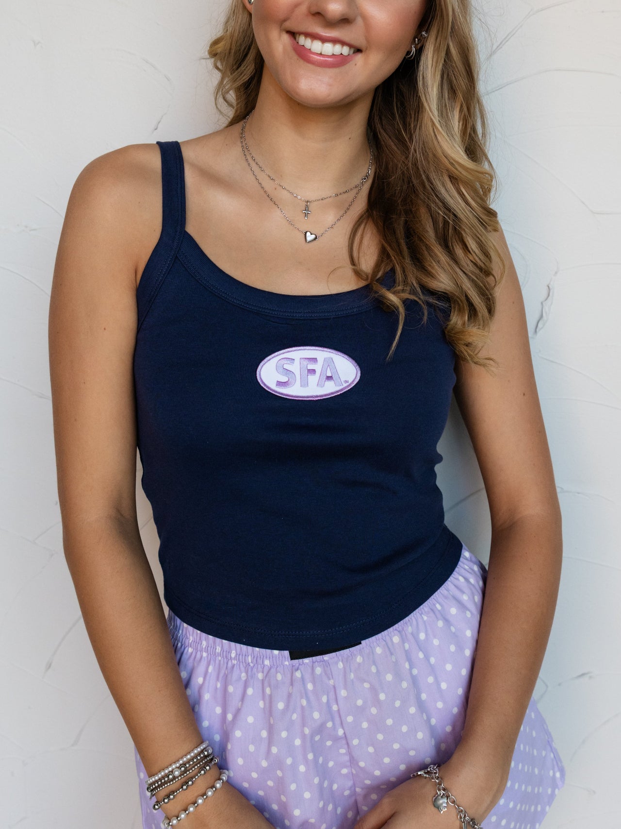 A mid-shot photograph focusing from the upper thighs to the mouth of a smiling young woman with wavy light brown hair posing against a textured white wall. She is wearing a fitted navy blue ribbed tank top made from 100% cotton fabric, featuring a scoop neckline and an oval purple embroidered patch centered at the chest reading "SFA", styled with lavender polka dot boxer shorts, layered silver necklaces, and stacked silver bracelets.