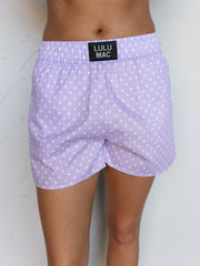 A close-up photograph focusing from the mid-thigh to the waist of a person standing against a textured white wall. They are wearing light purple lavender boxer shorts with a white polka dot pattern made from 100% cotton fabric, featuring a thick elastic waistband with a small rectangular black patch centered at the front that reads "LULU MAC".