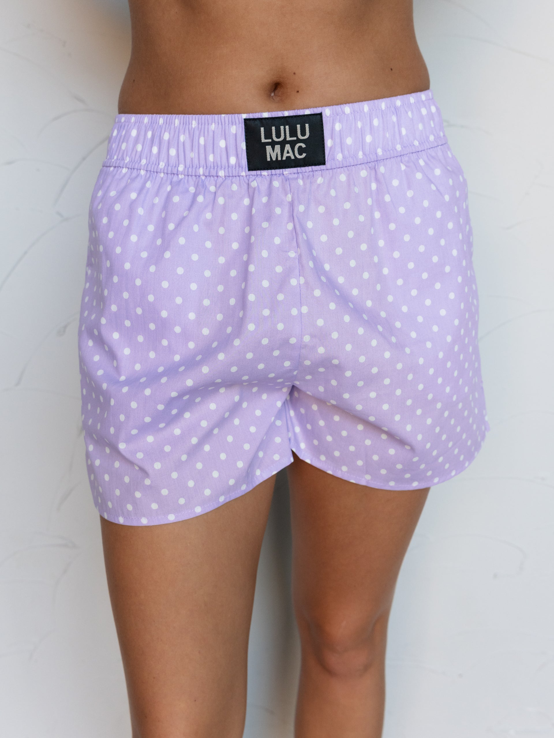 A close-up photograph focusing from the mid-thigh to the waist of a person standing against a textured white wall. They are wearing light purple lavender boxer shorts with a white polka dot pattern made from 100% cotton fabric, featuring a thick elastic waistband with a small rectangular black patch centered at the front that reads "LULU MAC".