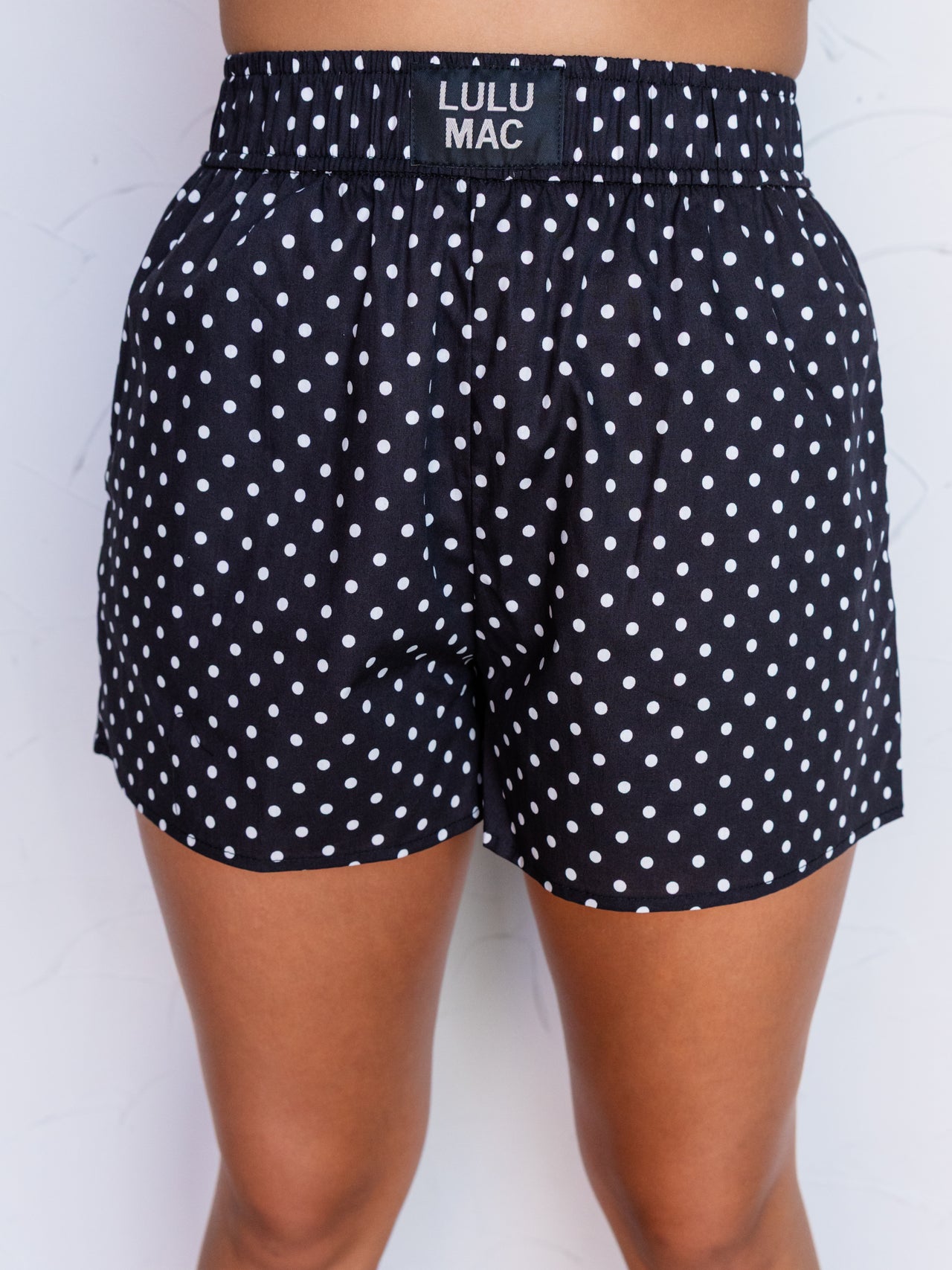 A close-up photograph focusing from the mid-thigh to the waist of a person standing against a textured white wall. They are wearing black boxer shorts with a white polka dot pattern made from 100% cotton fabric, featuring a thick elastic waistband with a small rectangular black patch centered at the front that reads "LULU MAC".