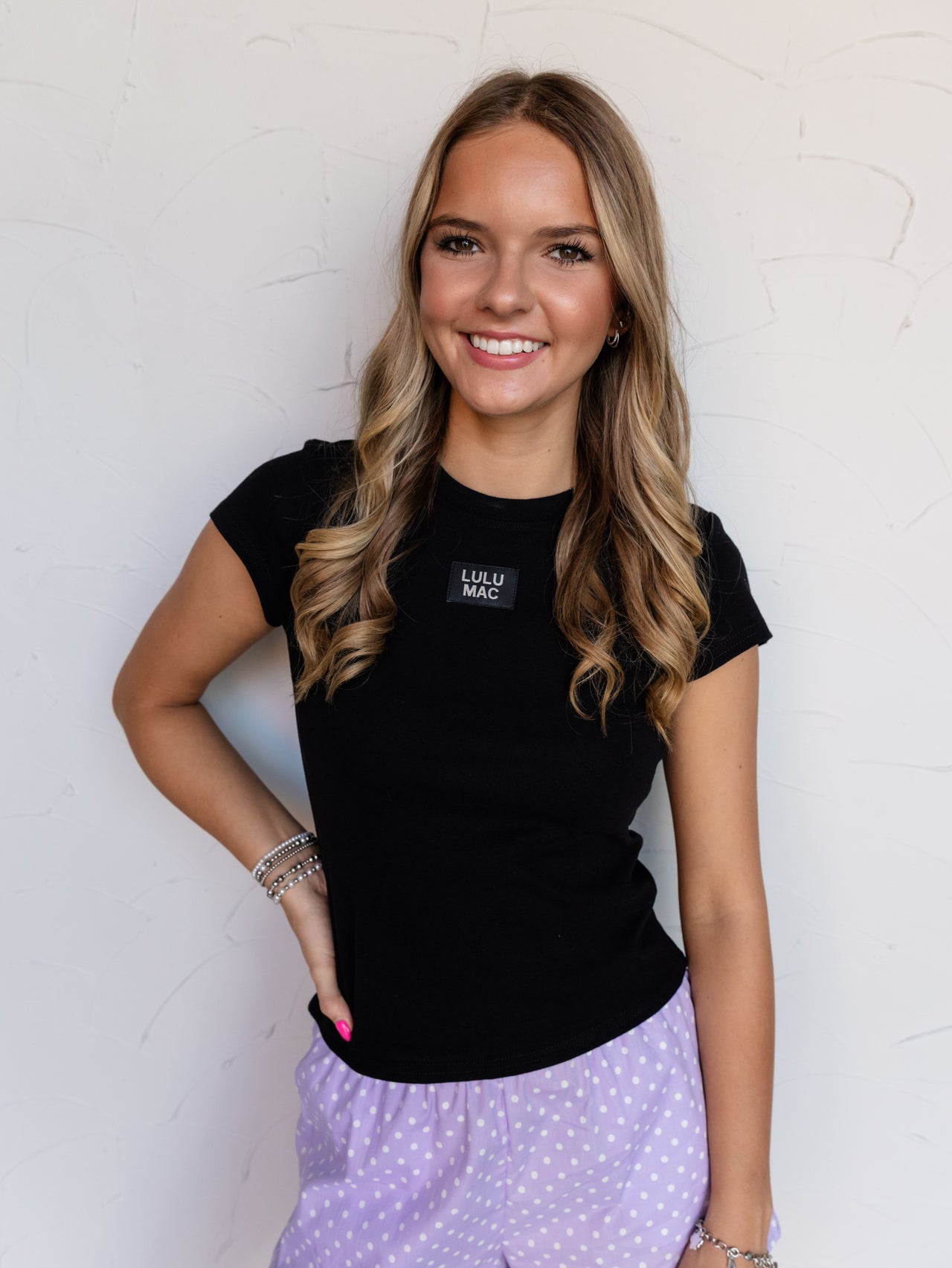 A mid-shot photograph focusing from the waist to the head of a smiling young woman with wavy light brown hair posing with one hand on her hip against a textured white wall. She is wearing a fitted solid black baby tee made from a 97% cotton and 3% spandex blend fabric, featuring a classic crew neckline and a small rectangular black patch at the center chest that reads "LULU MAC", styled with purple polka dot boxer shorts and stacked silver bracelets.