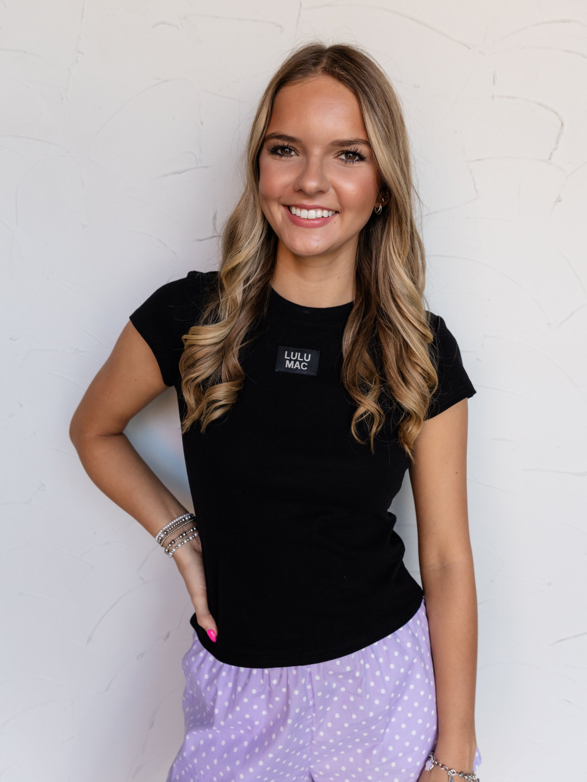 A mid-shot photograph focusing from the waist to the head of a smiling young woman with wavy light brown hair posing with one hand on her hip against a textured white wall. She is wearing a fitted solid black baby tee made from a 97% cotton and 3% spandex blend fabric, featuring a classic crew neckline and a small rectangular black patch at the center chest that reads "LULU MAC", styled with purple polka dot boxer shorts and stacked silver bracelets.