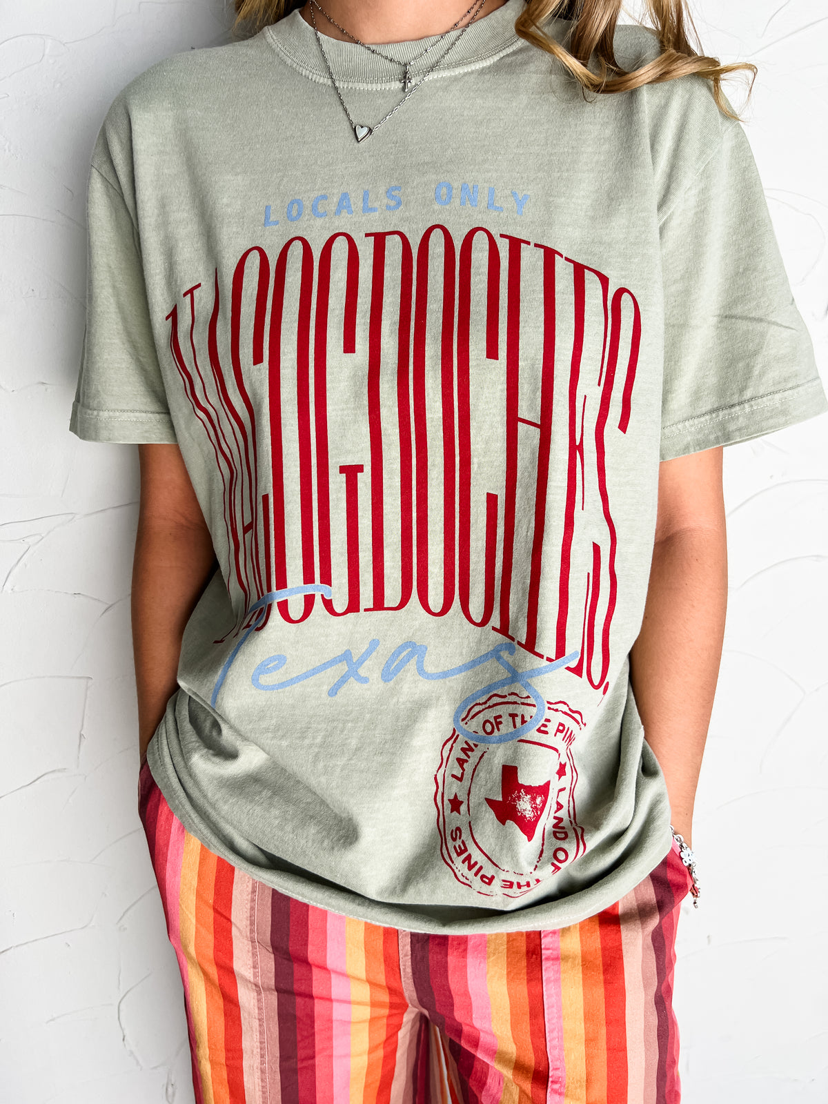 Catie wearing the Locals Only Nacogdoches Moss green Comfort Colors 100% ring-spun cotton tee in size Medium, featuring tall red Nacogdoches lettering, light blue Texas script, and a red Land of the Pines emblem, styled with colorful striped pants and layered necklaces.