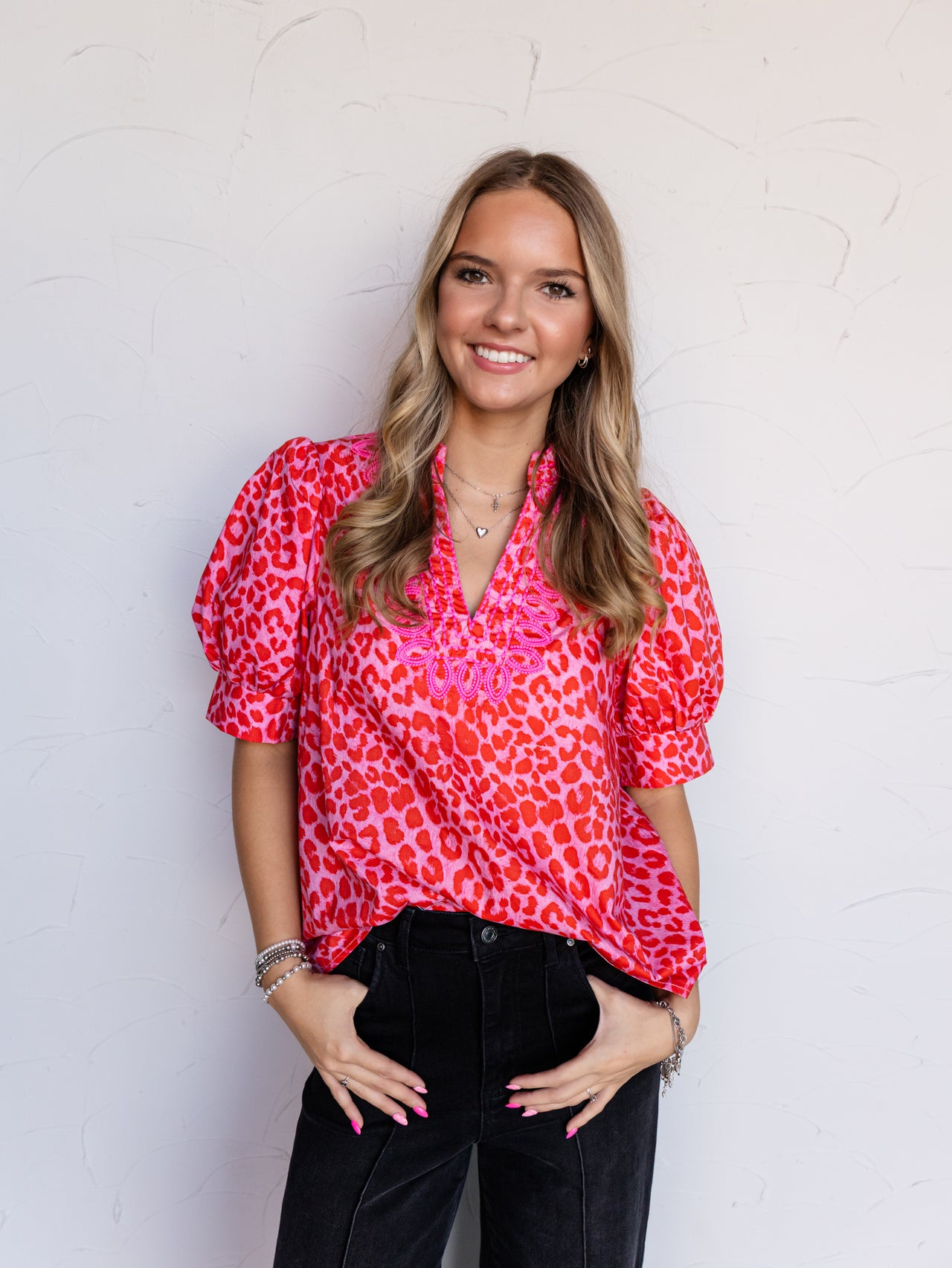 A portrait photograph focusing from the hips up of a smiling young woman with light brown wavy hair posing with her hands in her pockets against a textured white wall. She is wearing a short-sleeved blouse featuring a vibrant red and hot pink leopard print made from 100% cotton fabric, designed with a V-neckline adorned with pink embroidery and puff sleeves, styled with black denim jeans, layered silver necklaces, and stacked silver bracelets.