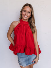A portrait photograph focusing from the hips up of a smiling young woman with light brown wavy hair posing against a textured white wall. She is wearing a sleeveless bright red high-neck halter top made from a 60% viscose and 40% polyester blend fabric, featuring a gathered neckline and a flared ruffled peplum hem, paired with a distressed denim mini skirt and silver bracelets.