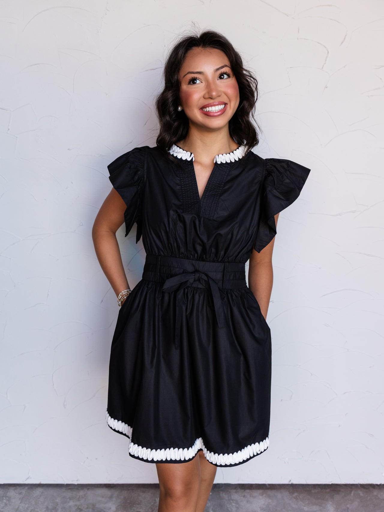 A portrait photograph focusing from the knees up of a smiling young woman with dark wavy hair looking off to the side against a textured white wall. She is wearing a black short-sleeved mini dress made from an 80% polyester and 20% cotton blend fabric, featuring flutter sleeves, a split V-neckline with white ric-rac trim, a tied waist belt, and matching white ric-rac trim at the hem, styled with stacked bracelets.
