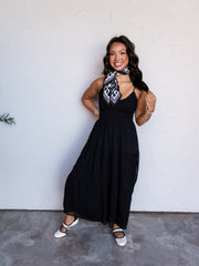 A full-length photograph of a smiling young woman with long dark wavy hair posing with one hand on her hip and the other touching her hair against a textured white wall. She is wearing a sleeveless black tiered maxi dress made from 100% polyester fabric, featuring a fitted smocked bodice and thin shoulder straps, styled with a black and white patterned neck scarf, white Mary Jane flats with black trim, and gold bracelets.