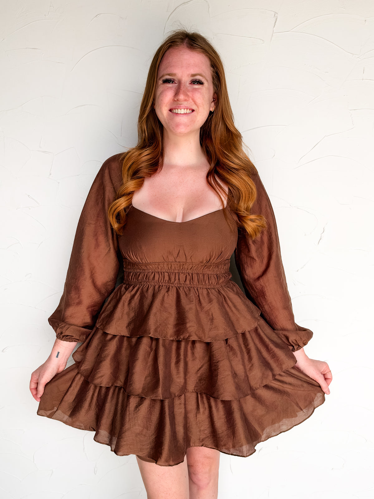 A smiling woman with long wavy light brown hair wearing a brown tiered mini dress with a smocked waist and sheer long sleeves, posing against a white textured wall.