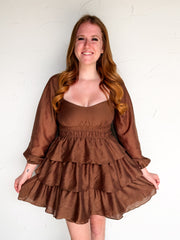 A smiling woman with long wavy light brown hair wearing a brown tiered mini dress with a smocked waist and sheer long sleeves, posing against a white textured wall.
