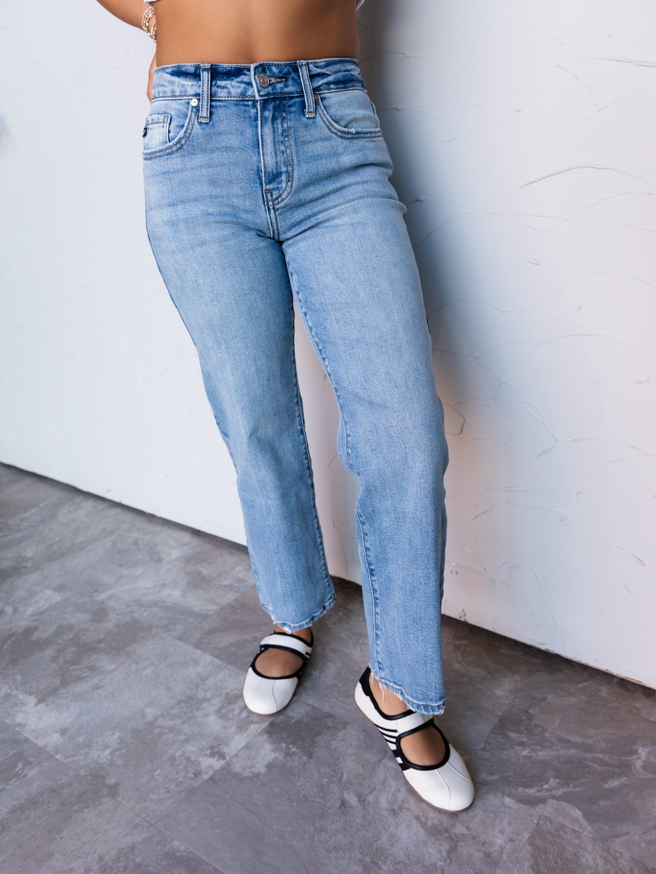 A photograph focusing from the waist down of a person leaning against a textured white wall. They are wearing medium-wash straight-leg jeans made from 100% cotton fabric, featuring a high-rise waist, classic five-pocket construction, belt loops, and slightly frayed ankle hems, styled with white Mary Jane flats with black straps.