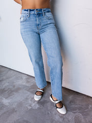 A photograph focusing from the waist down of a person leaning against a textured white wall. They are wearing medium-wash straight-leg jeans made from 100% cotton fabric, featuring a high-rise waist, classic five-pocket construction, belt loops, and slightly frayed ankle hems, styled with white Mary Jane flats with black straps.