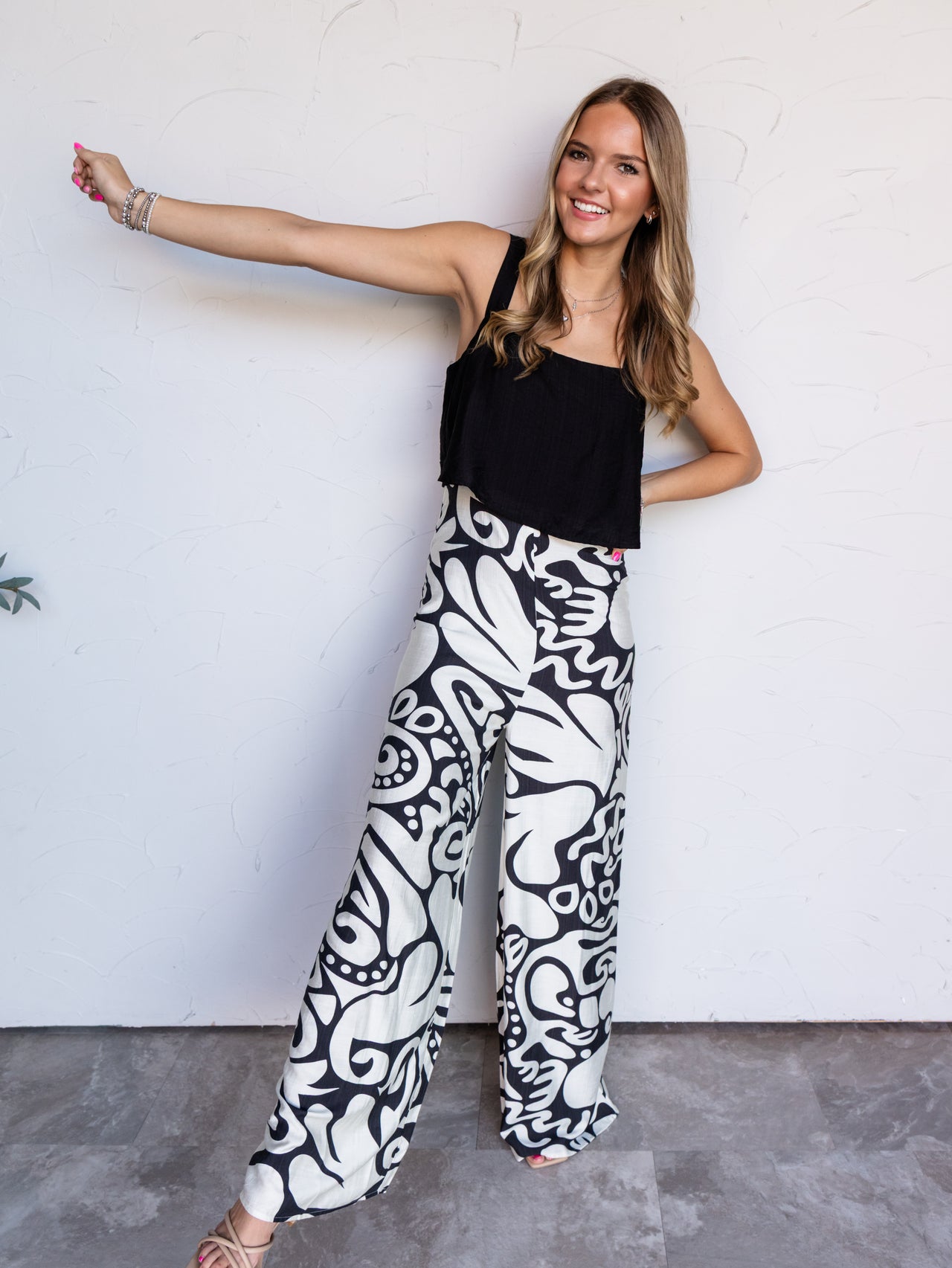 A full-length photograph of a smiling young woman with wavy light brown hair posing dynamically with one arm extended and the other on her hip against a textured white wall. She is wearing high-waisted wide-leg pants featuring a bold black and cream-white abstract swirl pattern, made from an 85% rayon and 15% polyamide blend fabric, styled with a solid black cropped tank top, nude strappy heels, and silver bracelets.