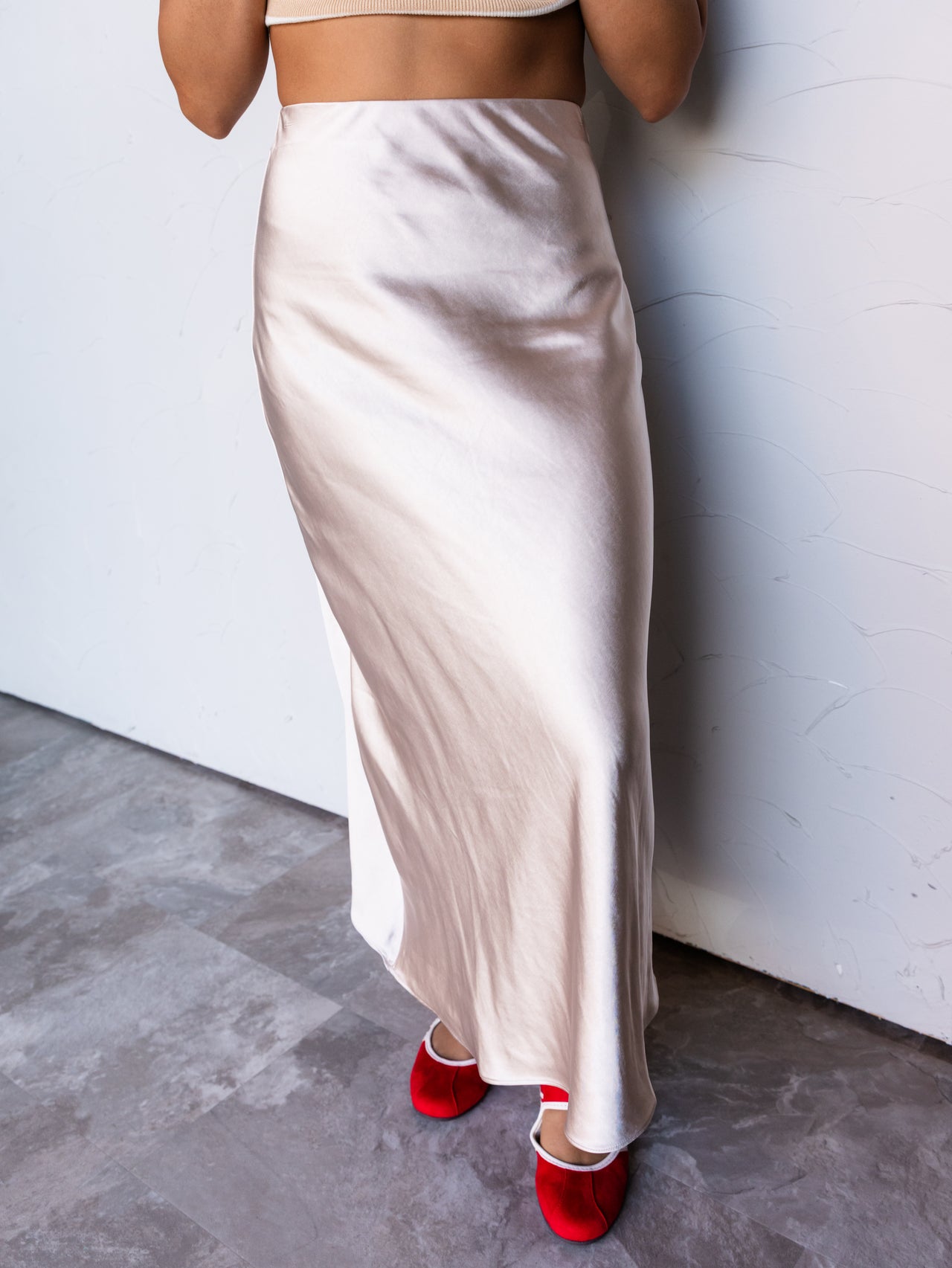A photograph focusing from the waist down of a person standing against a textured white wall. They are wearing a high-waisted champagne beige satin midi skirt made from 100% polyester fabric, featuring a smooth, glossy finish and a bias cut, styled with a tan ribbed cropped top and bright red velvet Mary Jane flats.