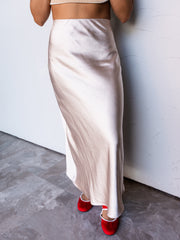 A photograph focusing from the waist down of a person standing against a textured white wall. They are wearing a high-waisted champagne beige satin midi skirt made from 100% polyester fabric, featuring a smooth, glossy finish and a bias cut, styled with a tan ribbed cropped top and bright red velvet Mary Jane flats.