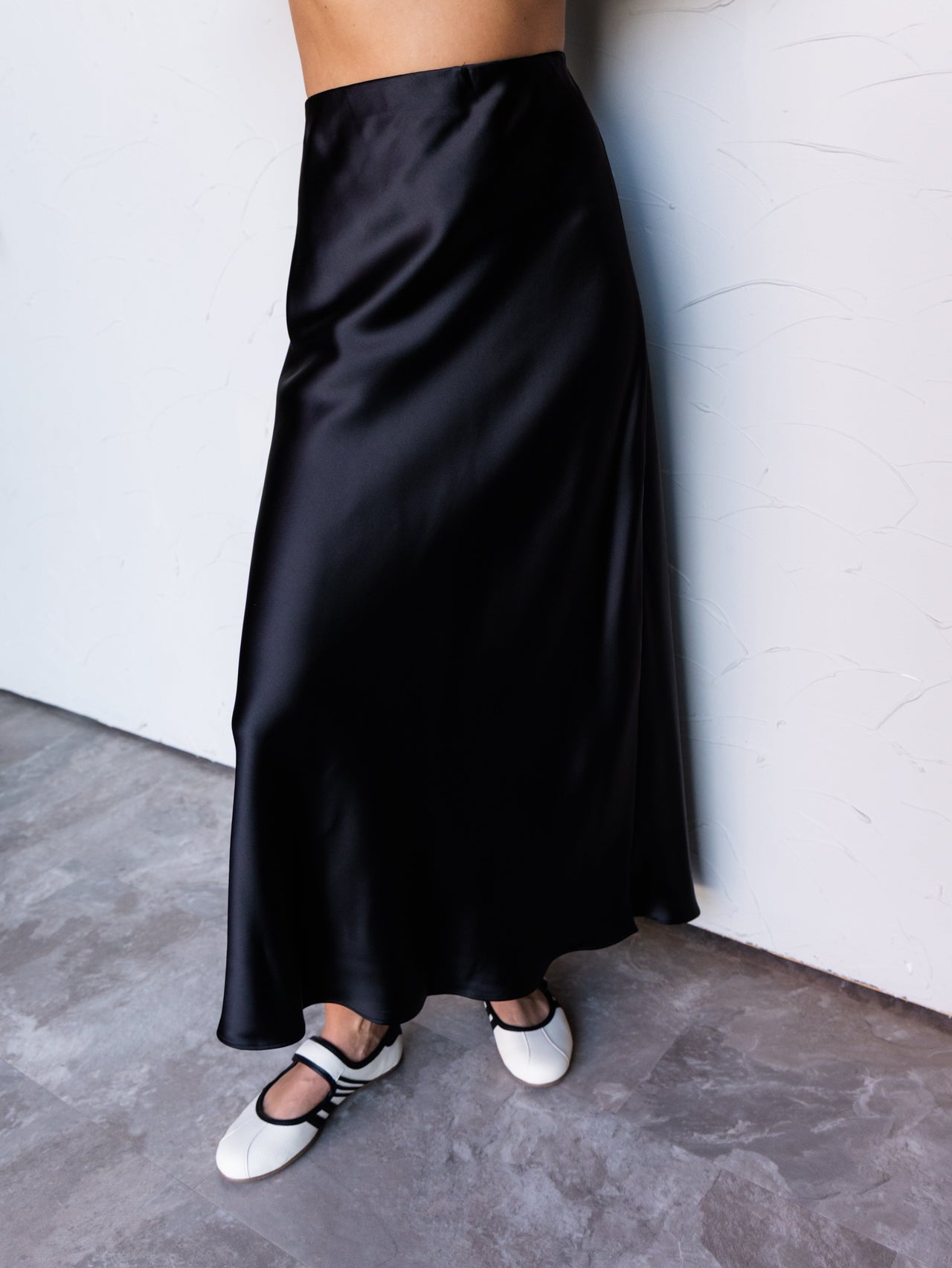 A photograph focusing from the lower waist to the feet of a person leaning against a textured white wall. They are wearing a high-waisted black satin midi skirt made from 100% polyester fabric, featuring a glossy sheen and a flowing bias cut, styled with a black-and-white ribbed crop top and white Mary Jane flats with black straps.