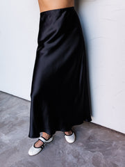 A photograph focusing from the lower waist to the feet of a person leaning against a textured white wall. They are wearing a high-waisted black satin midi skirt made from 100% polyester fabric, featuring a glossy sheen and a flowing bias cut, styled with a black-and-white ribbed crop top and white Mary Jane flats with black straps.