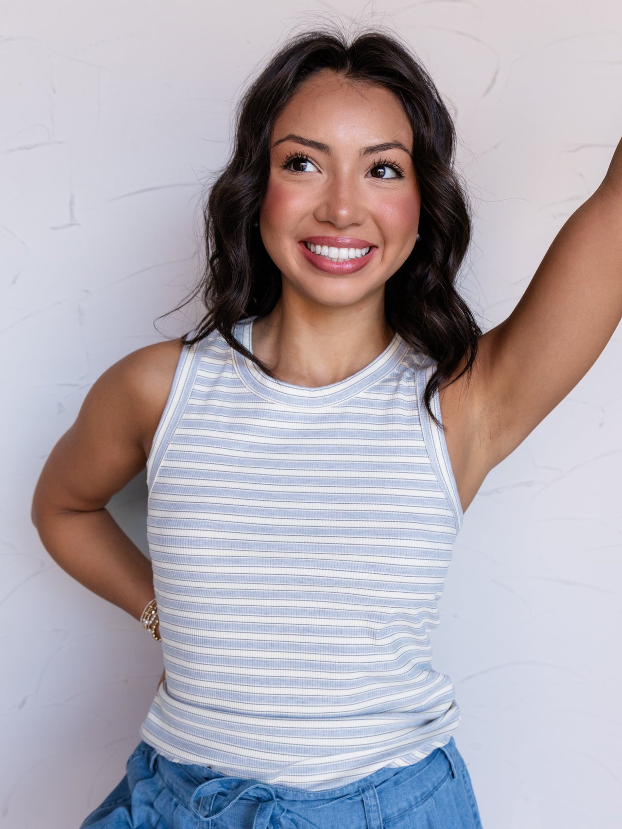 A portrait photograph focusing from the waist up of a smiling woman with shoulder-length wavy dark hair posing with one arm raised against a textured white wall. She is wearing a sleeveless ribbed tank top with thin light blue and cream horizontal stripes made from a 95% polyester and 5% spandex blend fabric, featuring a crew neck and rounded hemline, styled with chambray ruffled drawstring shorts and a bracelet.