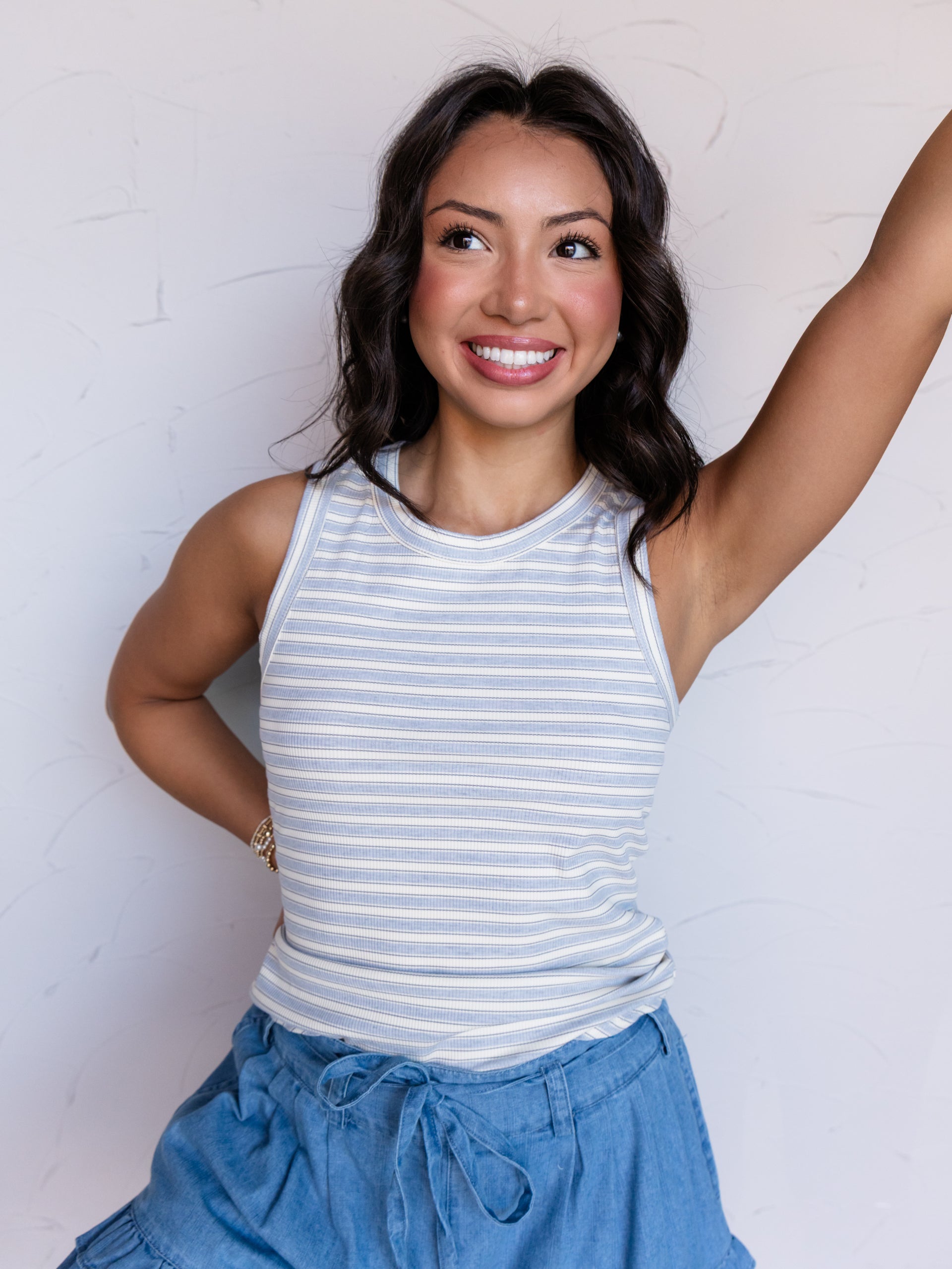 A portrait photograph focusing from the waist up of a smiling woman with shoulder-length wavy dark hair posing with one arm raised against a textured white wall. She is wearing a sleeveless ribbed tank top with thin light blue and cream horizontal stripes made from a 95% polyester and 5% spandex blend fabric, featuring a crew neck and rounded hemline, styled with chambray ruffled drawstring shorts and a bracelet.