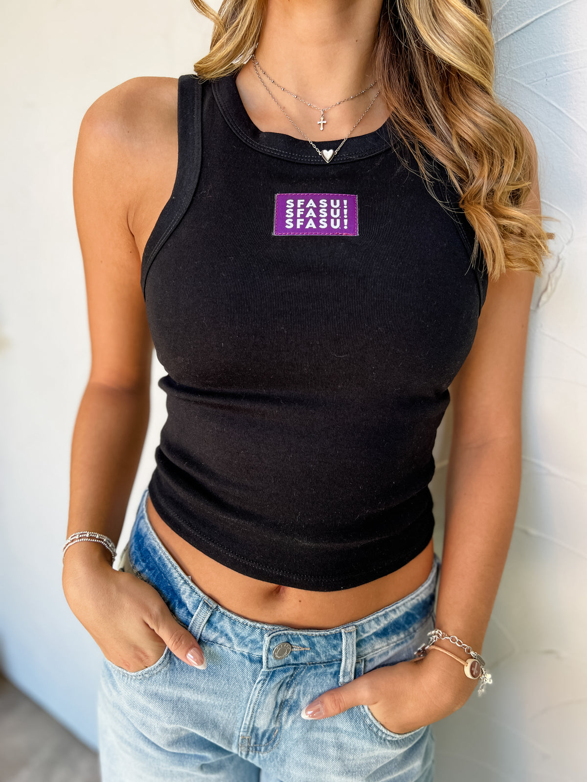 Model wearing a black SFASU ribbed racer tank by G Ink, made of 52% cotton and 48% polyester, featuring a high rounded neckline and purple chest patch with white SFASU lettering, paired with light-wash jeans.