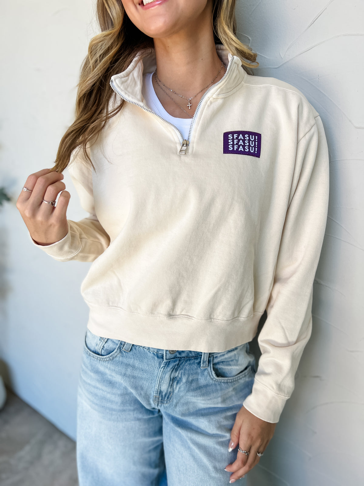 Model wearing an ivory garment-dyed SFASU quarter-zip sweatshirt by G Ink, made of 80% cotton and 20% polyester, with a stand collar, long sleeves, banded hem, and purple chest patch with white SFASU lettering, paired with light-wash jeans.