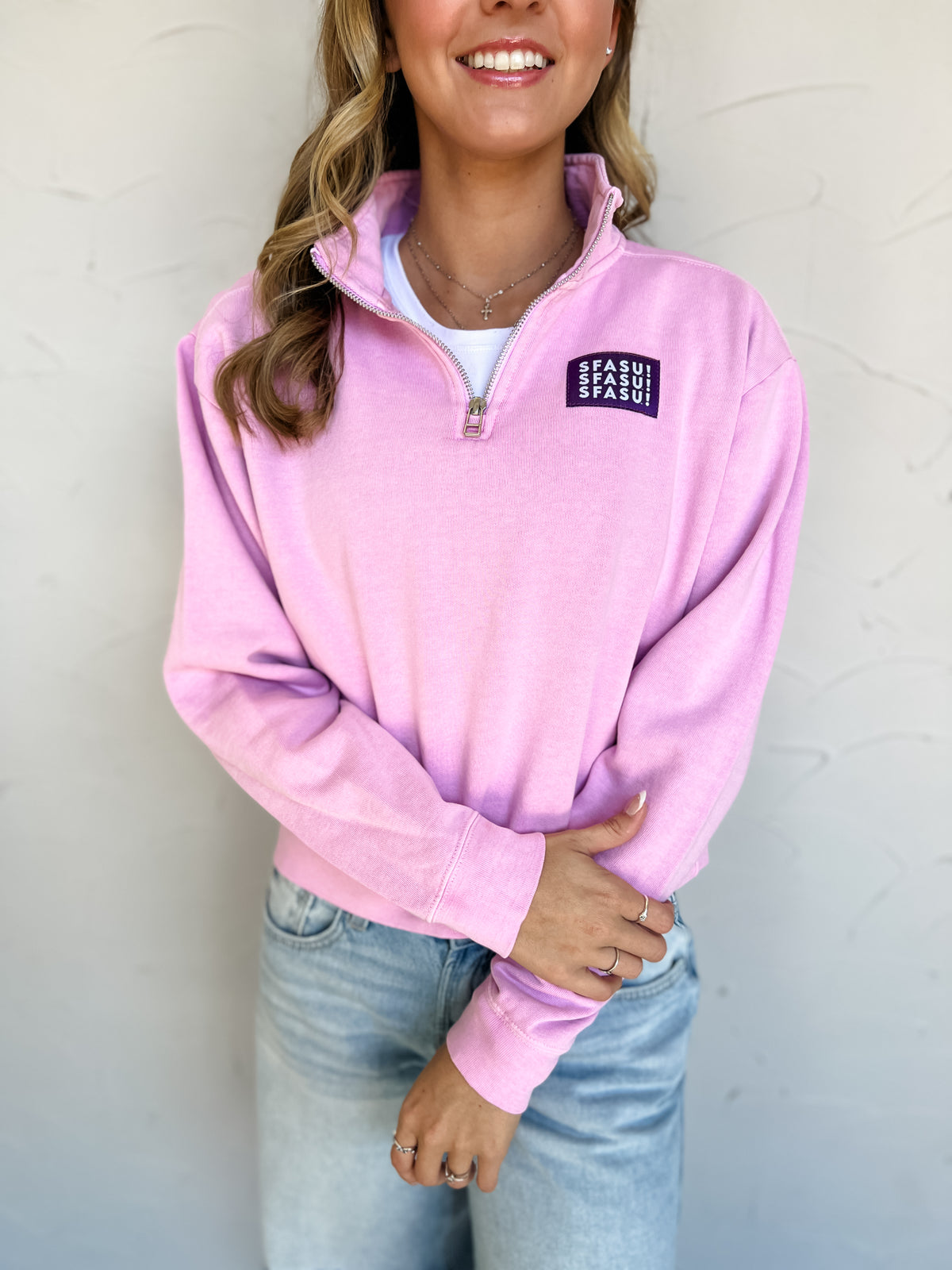 Model wearing a pink garment-dyed SFASU quarter-zip sweatshirt by G Ink, made of 80% cotton and 20% polyester, with a stand collar, long sleeves, banded cuffs and hem, and a purple chest patch with white SFASU lettering, paired with light-wash jeans.