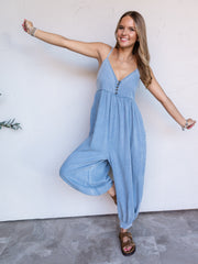 A full-length photograph of a smiling woman with long blonde hair with dark roots posing playfully with arms stretched out and one leg raised against a textured white wall. She is wearing a soft light-wash denim jumpsuit made from a 65% cotton and 35% Tencel blend fabric, featuring thin shoulder straps, a V-neckline with decorative buttons, a wide slouchy drop-crotch fit, and elasticized ankle hems, styled with brown buckled slide sandals and layered silver bracelets.