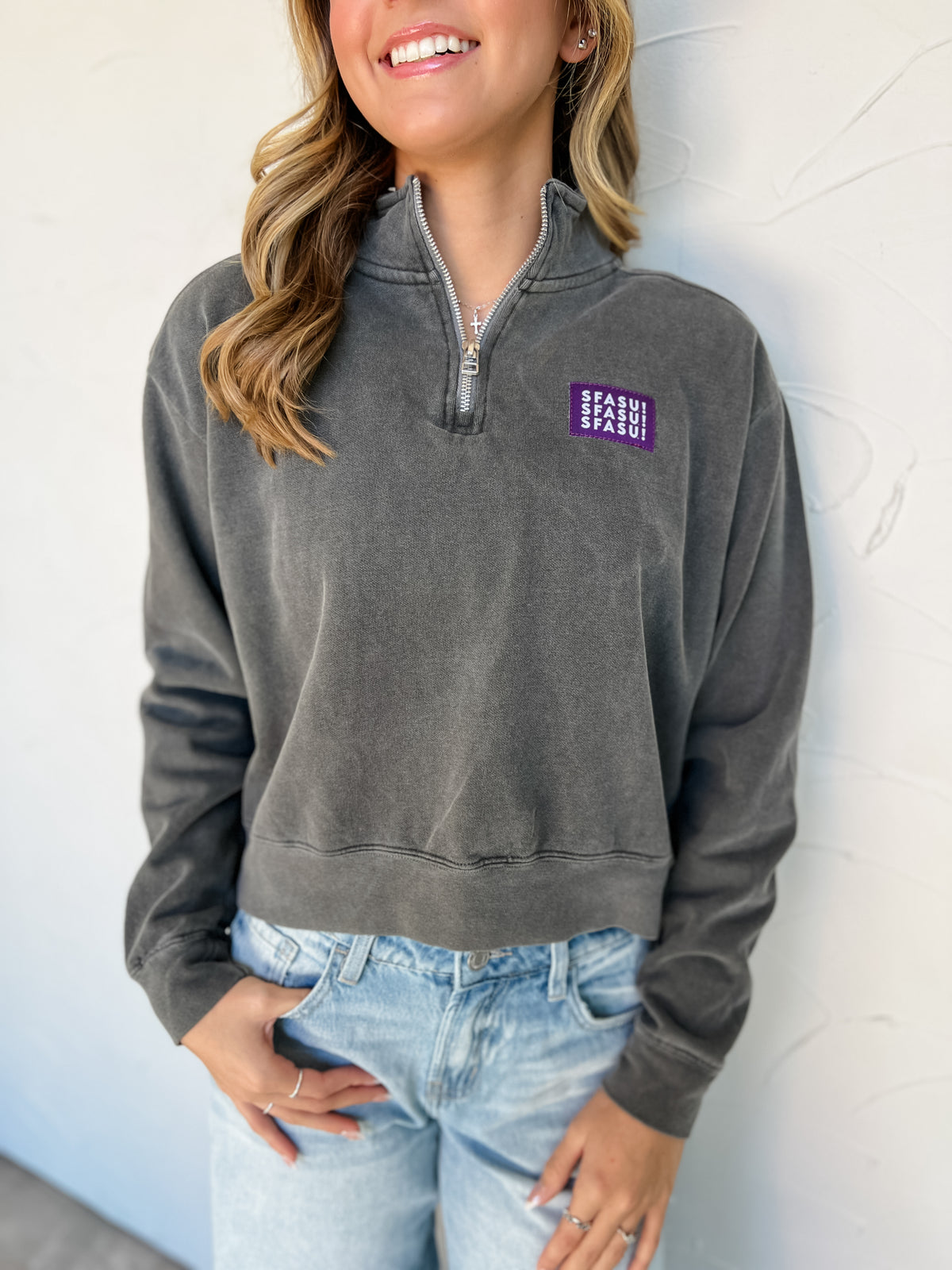 Model wearing a Pepper charcoal-gray garment-dyed SFASU quarter-zip sweatshirt by G Ink, made of 80% cotton and 20% polyester, with a stand collar, long sleeves, banded cuffs and hem, and a purple chest patch with white SFASU lettering, paired with light-wash jeans.