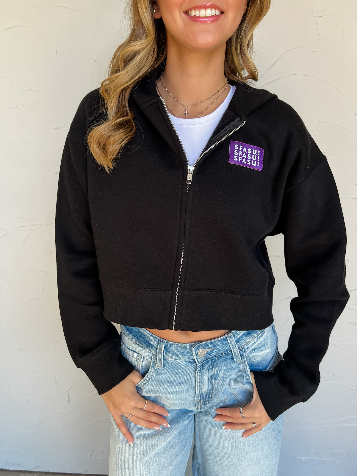 Model wearing a black SFASU cropped full-zip hoodie by G Ink, made of 80% combed cotton and 20% recycled polyester, with the hood up, long sleeves, banded cuffs and hem, and a purple chest patch with white SFASU lettering, paired with light-wash jeans.