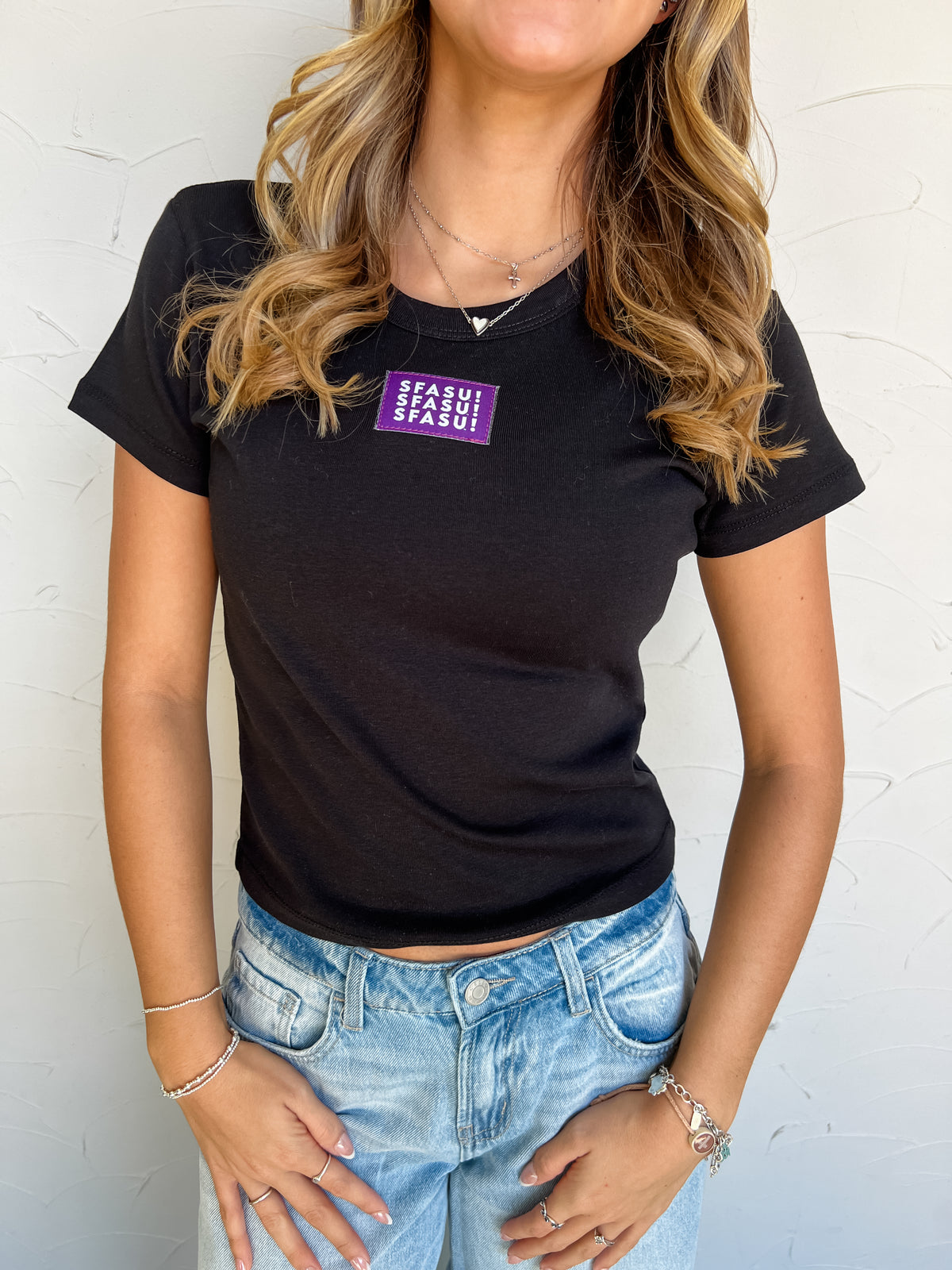 Model wearing a black 100% cotton SFASU baby tee by G Ink, featuring a rounded neckline, short sleeves, fitted silhouette, and a purple chest patch with white SFASU lettering, paired with light-wash jeans.