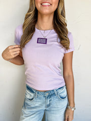 Model wearing a lilac 100% cotton SFASU baby tee by G Ink, featuring a rounded neckline, short sleeves, fitted silhouette, and a purple chest patch with white SFASU lettering, paired with light-wash jeans.
