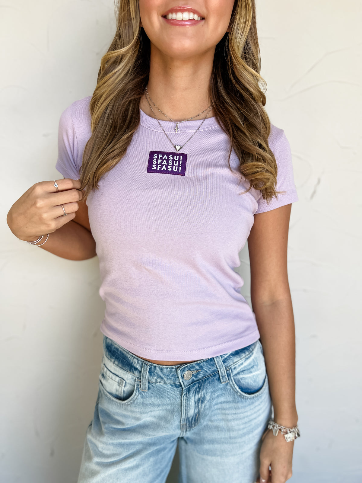 Model wearing a lilac 100% cotton SFASU baby tee by G Ink, featuring a rounded neckline, short sleeves, fitted silhouette, and a purple chest patch with white SFASU lettering, paired with light-wash jeans.