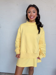 A three-quarter length photograph of a smiling woman with long dark wavy hair posing against a textured white wall. She is wearing a matching butter yellow two-piece fleece lounge set made from an 80% cotton and 20% polyester blend, featuring an oversized long-sleeve mock neck pullover sweatshirt with dropped shoulders and ribbed hem band, paired with a high-waisted elastic drawstring mini skirt, styled with pearl stud earrings.