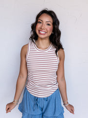 A portrait photograph focusing from the waist up of a smiling woman with shoulder-length wavy dark hair posing against a textured white wall. She is wearing a sleeveless ribbed tank top with thin mauve and cream horizontal stripes made from a 95% polyester and 5% spandex blend fabric, featuring a crew neck and rounded hemline, styled with chambray ruffled drawstring shorts and beaded stack bracelets.