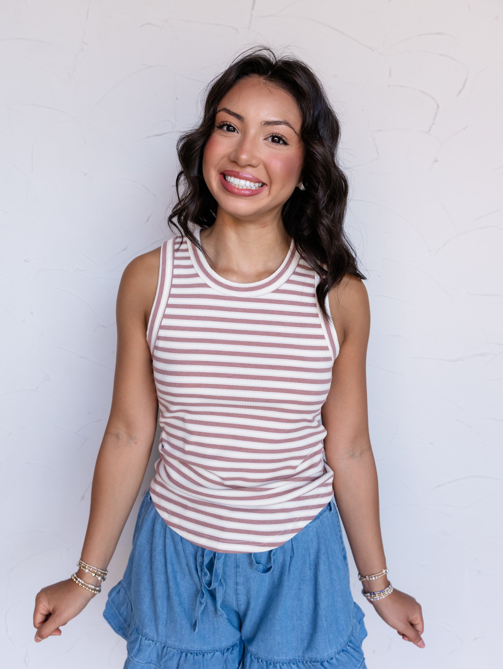 A portrait photograph focusing from the waist up of a smiling woman with shoulder-length wavy dark hair posing against a textured white wall. She is wearing a sleeveless ribbed tank top with thin mauve and cream horizontal stripes made from a 95% polyester and 5% spandex blend fabric, featuring a crew neck and rounded hemline, styled with chambray ruffled drawstring shorts and beaded stack bracelets.