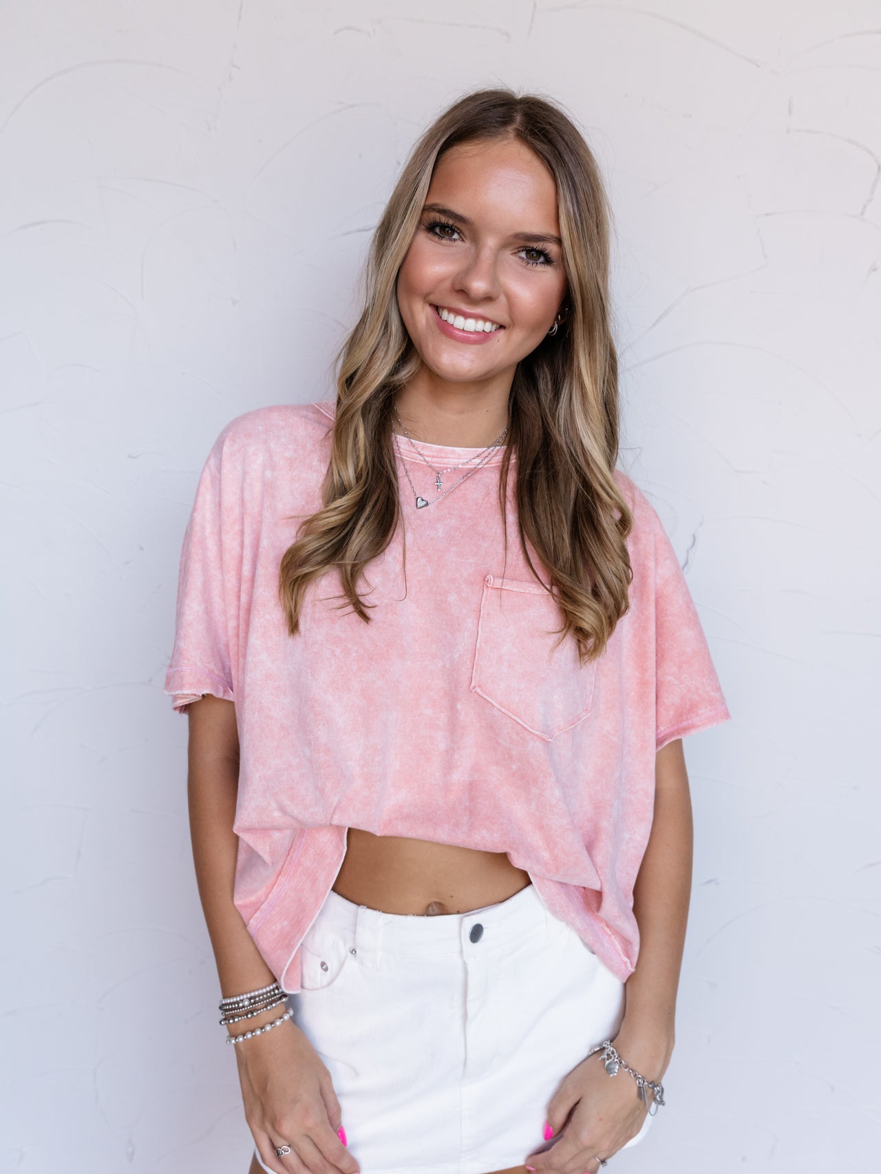 A portrait photograph focusing from the hips up of a smiling woman with long wavy blonde hair posing with her hands resting in front against a textured white wall. She is wearing an oversized pink mineral wash short-sleeved tee made from 100% cotton fabric, featuring a crew neckline, dropped shoulders, and a single chest patch pocket, styled with a high-waisted white denim miniskirt and layered silver necklaces and bracelets.